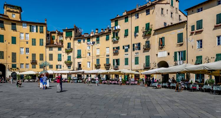 Une piazza ensoleillée et historique encadrée par des bâtiments jaunes aux volets verts et des parasols de café en plein air.