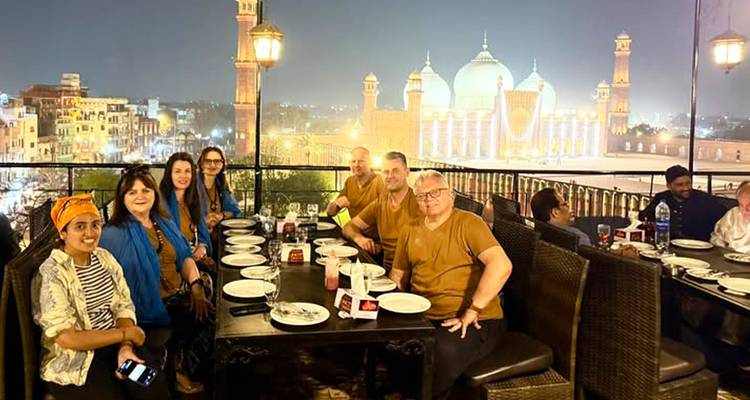 Large dinner gathering on a rooftop restaurant overlooking the illuminated Badshahi Mosque at night.