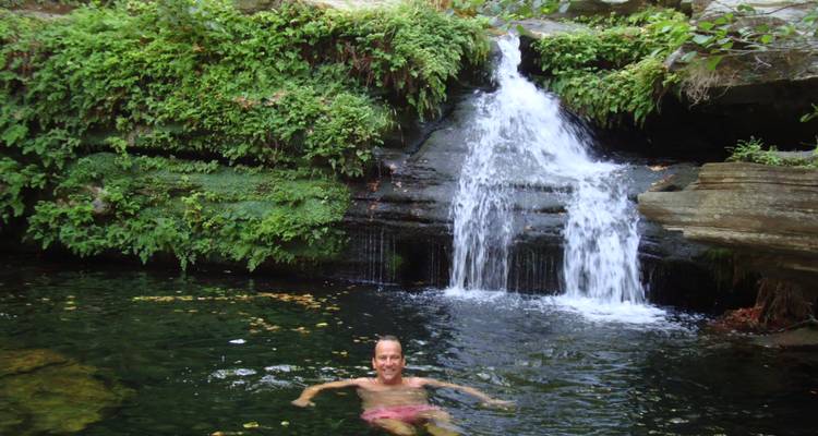 Une femme nage dans un bassin naturel limpide sous une petite cascade forestière bordée de fougères.