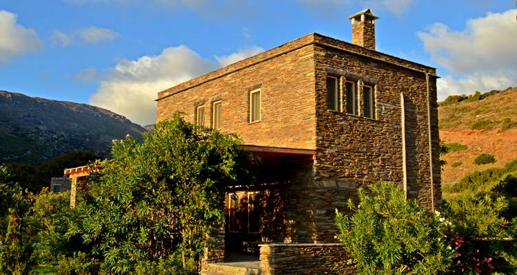 Villa en pierre rustique entourée d'arbustes avec des montagnes et un ciel bleu en arrière-plan.