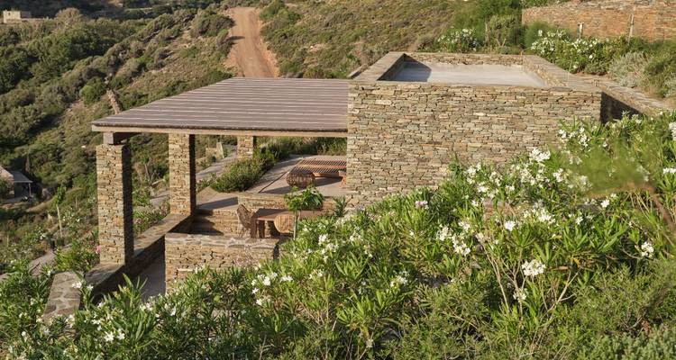 Terrasse en pierre avec pergola surplombe un sentier verdoyant sur une colline menant vers la mer Égée.