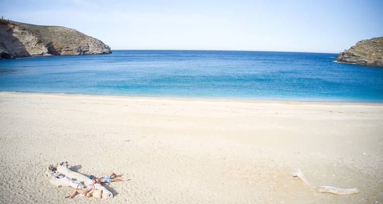 Vaste plage déserte avec une mer azur où deux personnes se détendent à côté d'un grand tronc d'arbre échoué.