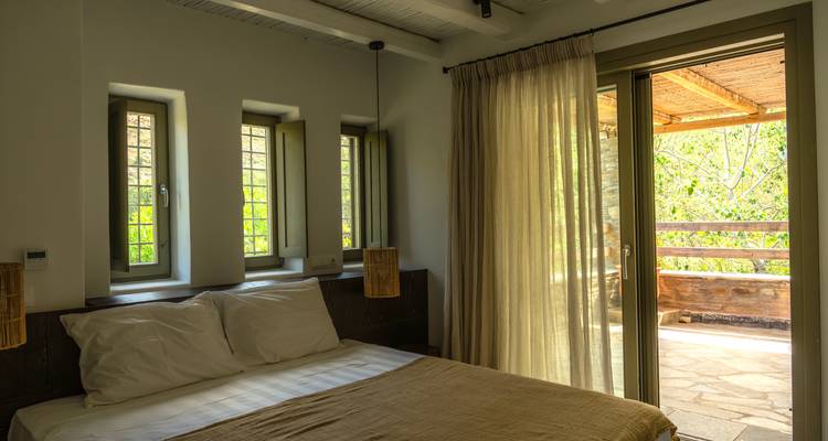 Chambre confortable avec linge de lit neutre, trois petites fenêtres et porte-fenêtre donnant sur un balcon avec rideaux.