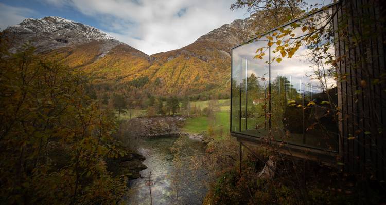 Cabaña moderna de vidrio ubicada sobre un desfiladero de río con montañas otoñales al fondo