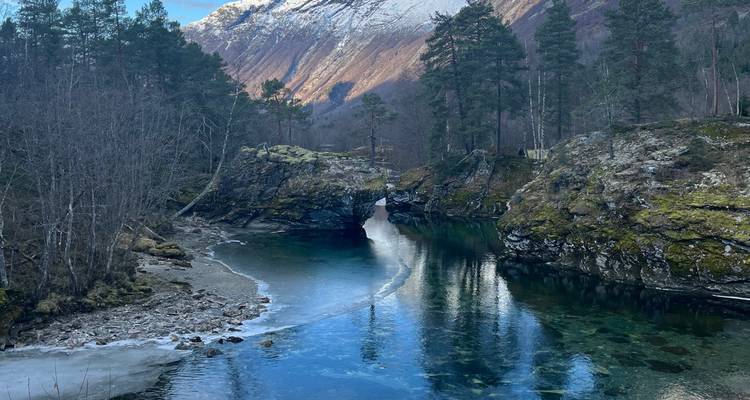 Río claro que fluye entre orillas rocosas con árboles desnudos y montañas cubiertas de nieve al fondo