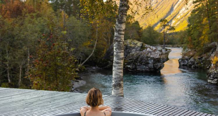 Persona remojándose en jacuzzi al aire libre en una terraza de madera con vista al río y al bosque dorado de otoño