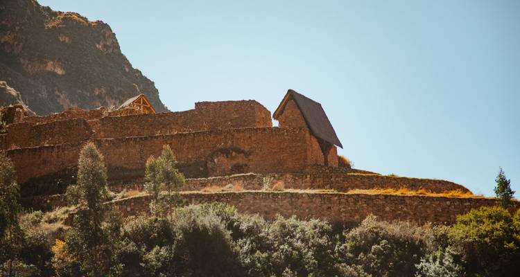 Stone Inca terraces and a thatched roof structure cling to a sunlit Andean hillside.