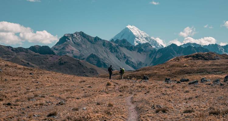 Two trekkers walk an open alpine plain with a snow-capped Andean peak dominating the skyline.