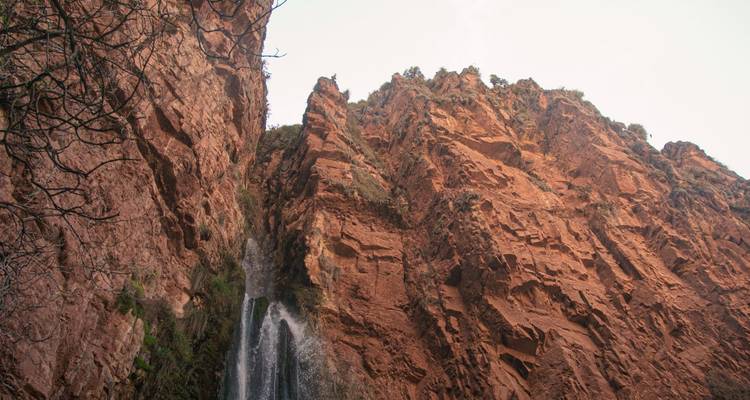 Water cascades from a hidden waterfall down a sheer red rock cliff against a bright sky.