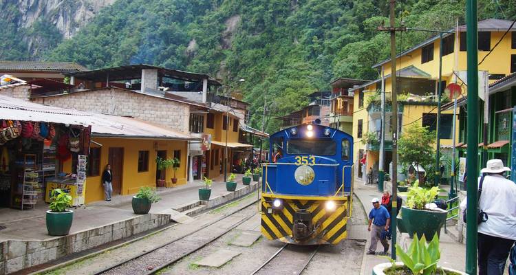 Ein blau-gelber Zug rollt durch die farbenfrohen Straßen von Aguas Calientes neben üppigen Dschungelhängen.
