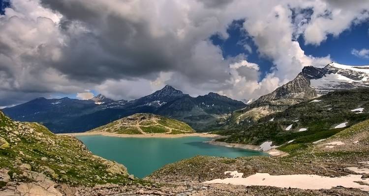 Ein türkisfarbener Alpensee liegt inmitten dramatischer, von Wolken gekrönter Schneiberge in unberührter Hochlandlandschaft.