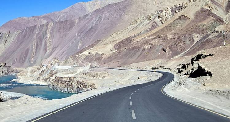 Eine glatte Asphaltstraße schlängelt sich durch karge Hochgebirgsmountains neben einem blauen Flussbett.
