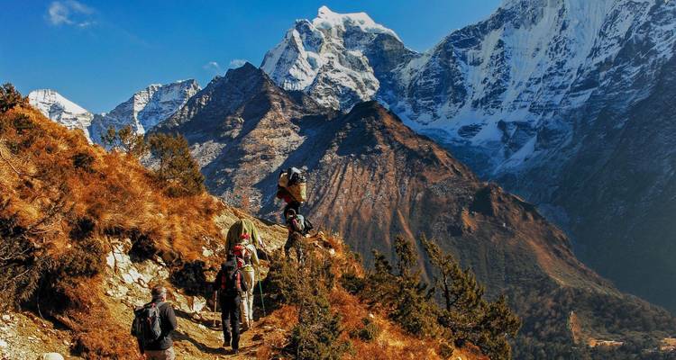 Rucksacktouristen wandern einen steilen Bergpfad hinauf zu hoch aufragenden schneebedeckten Gipfeln vor einem klaren Himmel.