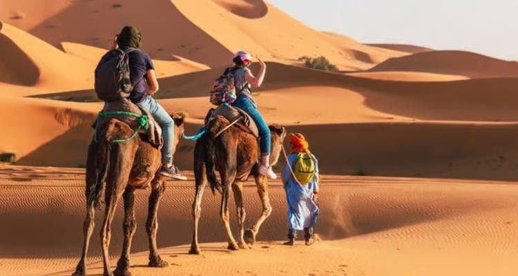 Turistas montando camellos guiados por un guía a través de dunas doradas del desierto.