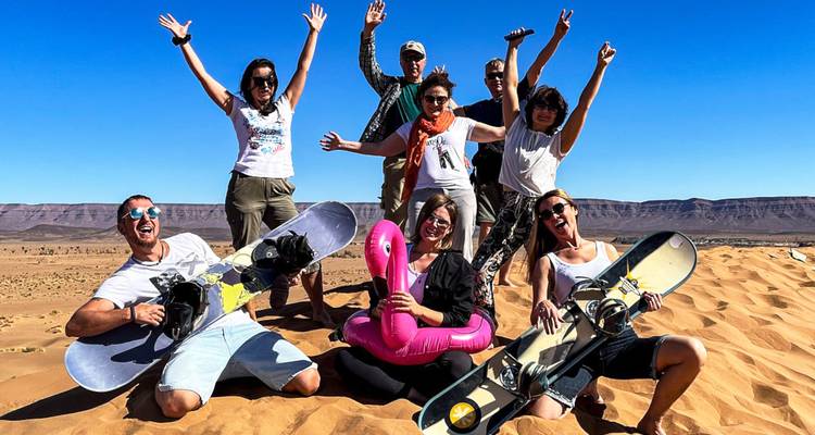 Grupo enérgico de viajeros sentados en dunas de arena sosteniendo tablas de snowboard y juguete inflable bajo un cielo azul despejado.