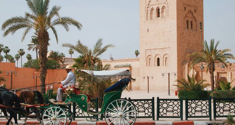 Carruaje tirado por caballos pasando entre palmeras frente a un alto minarete de arenisca en una ciudad marroquí.