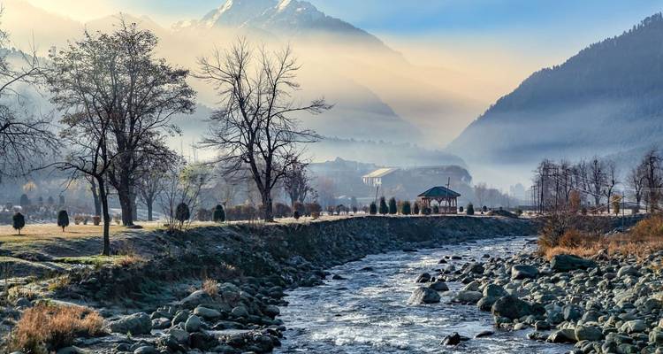 Un ruisseau de montagne limpide coule à travers une vallée givrée avec une brume matinale et un sommet enneigé au loin.