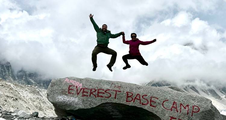 Two trekkers mid-air leap atop boulder marked 'Everest Base Camp'.