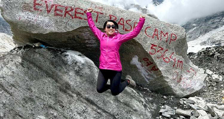 Female trekker jumps with joy beside engraved Everest Base Camp boulder.