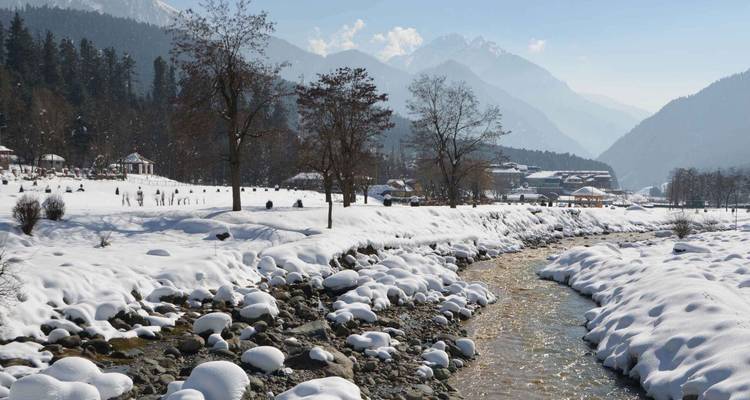 Schneebeladene Flussufer und Alpendorf unterhalb geschichteter Himalaya-Gebirgsketten an einem sonnigen Wintertag.