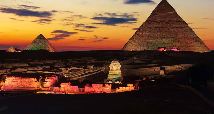 Vista nocturna de la Esfinge y las pirámides iluminadas con luces coloridas contra un cielo anaranjado del atardecer.