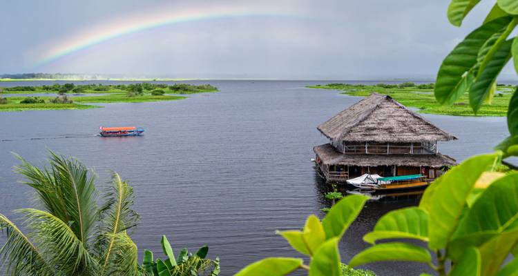 Lodge amazonien sur pilotis sous un arc-en-ciel enjambant une large rivière et une plaine inondable luxuriante