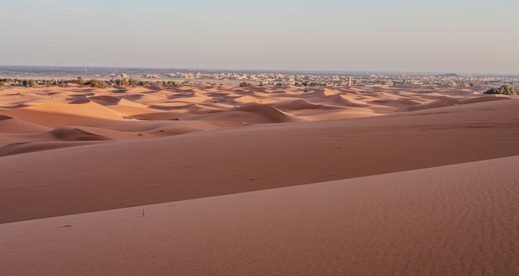 Des dunes de sable orange s'étendent majestueusement vers un village désertique lointain à l'horizon.
