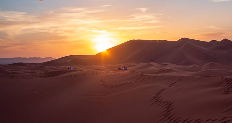 De zon zakt weg achter golvende zandduinen terwijl kleine groepjes op de rand zitten om naar de kleuren te kijken.