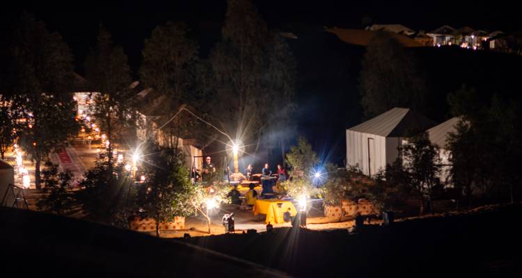 Nachtelijke woestijnkamp met gloeïende lantaarns, eettenten en gasten die rond tafels verzameld zijn.