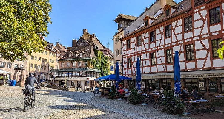 Geplaveide plein in Neurenberg omzoomd met vakwerkhuizen, buitenterrassen en een langsfietende wielrenner.