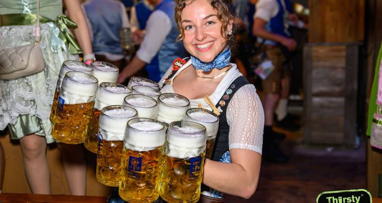 Glimlachende serveerster in dirndl die een hoge stapel schuimende bierpullen draagt in een feestelijke zaal.