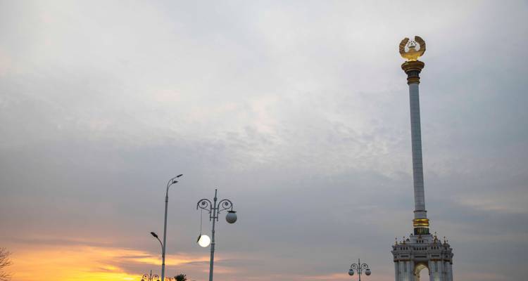 Une haute colonne portant un emblème doré s'élève contre un ciel nuageux au lever du soleil.