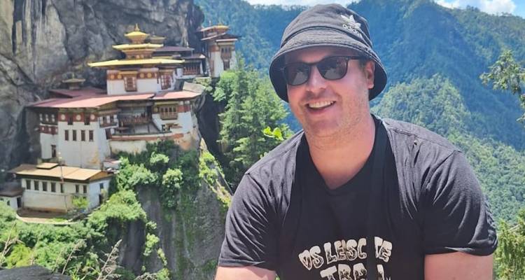 Traveler in dark hat smiling with Bhutan's cliffside monastery perched behind.
