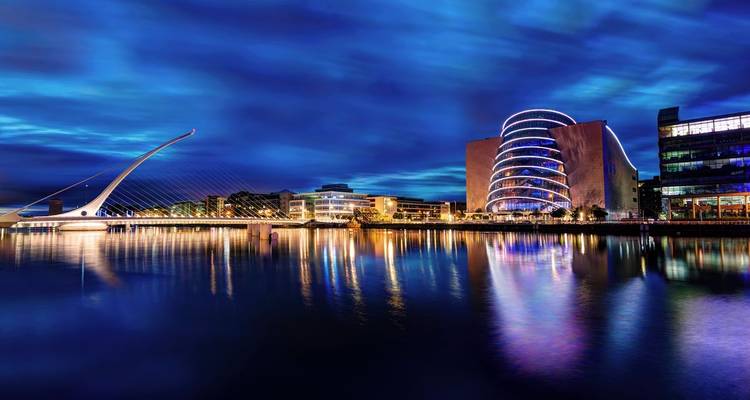 Night skyline of Dublin's riverfront with Samuel Beckett Bridge and modern Convention Centre reflecting in calm water.