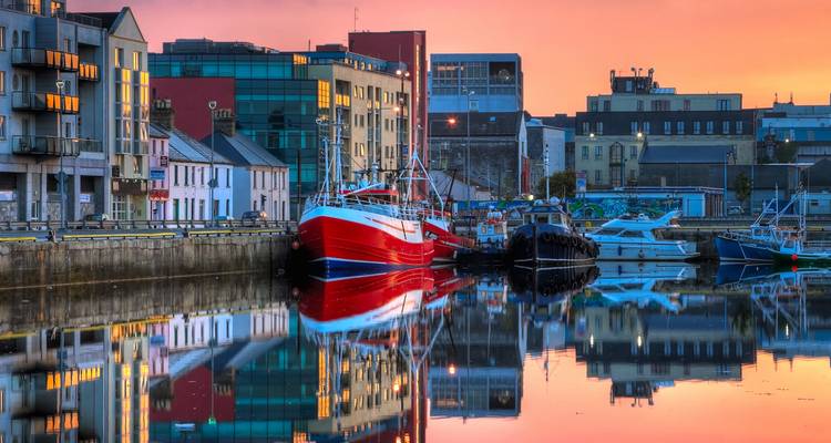 Colorful fishing boats and modern buildings reflect in calm water during a vibrant sunset in Galway.
