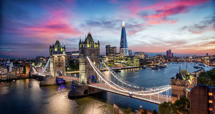 Dramatic twilight panorama of London's Tower Bridge and the Shard with streaks of city lights over the Thames.