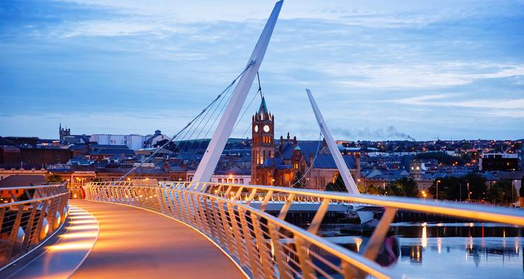 Modern arch pedestrian bridge curving over a calm river with a historic clock-tower cityscape at dusk, lights glowing along the rail.