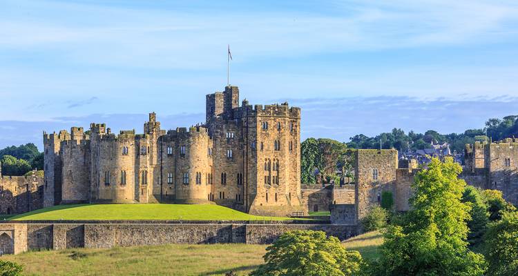 Large stone medieval castle set on a grassy mound under a clear blue sky, surrounded by trees and countryside.