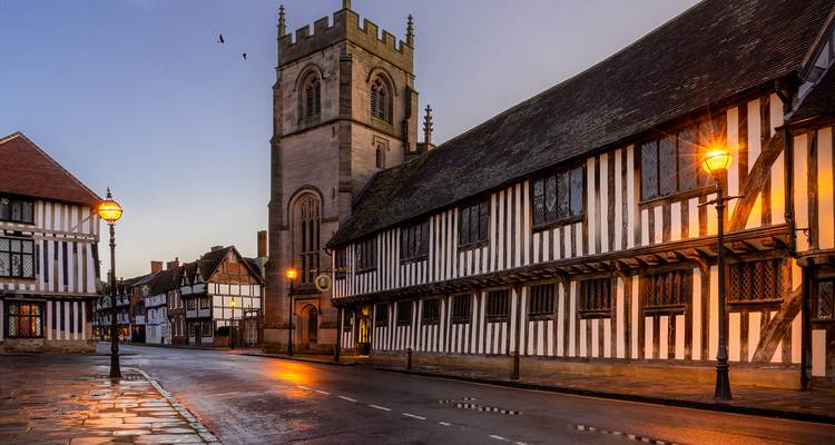 Quiet cobbled street lined with black-and-white Tudor buildings leading to a stone church tower in soft evening light.