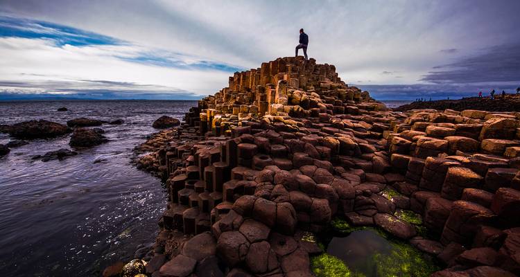 Dramatic hexagonal basalt columns rising from the Atlantic with a lone hiker standing on the summit under moody skies.