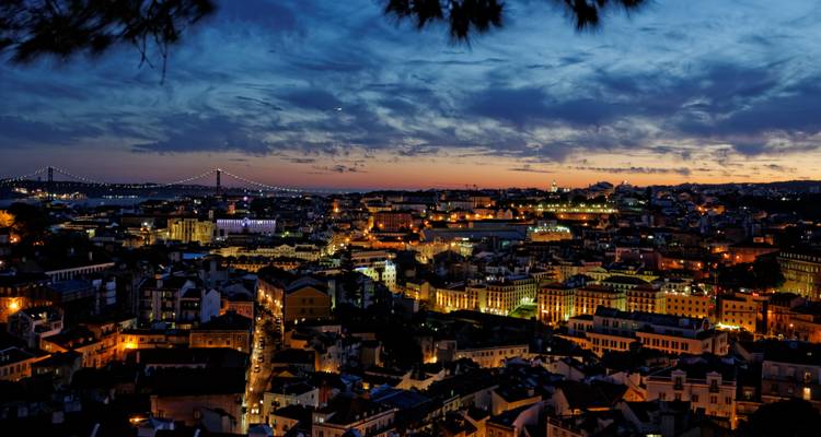 Panorama crepuscular de Lisboa con luces de la ciudad brillando, el Puente 25 de Abril y un cielo dramático azul-naranja.