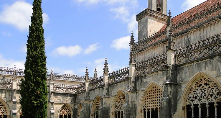 Claustro de piedra con arcos ornamentados y agujas que rodean un jardín de patio bajo el cielo azul.