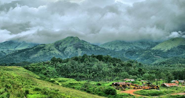 Weelderige beboste heuvels en bewolkte lucht boven het landelijke Wayanad landschap met kleine gebouwen eronder.