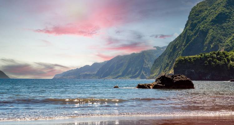 Las tranquilas aguas del Atlántico acarician una playa aislada de Madeira respaldada por exuberantes acantilados verdes bajo un cielo teñido de rosa.