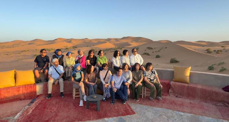 Grote tourgroep poseert op kussens met uitzicht op zandduinen bij zonsondergang.