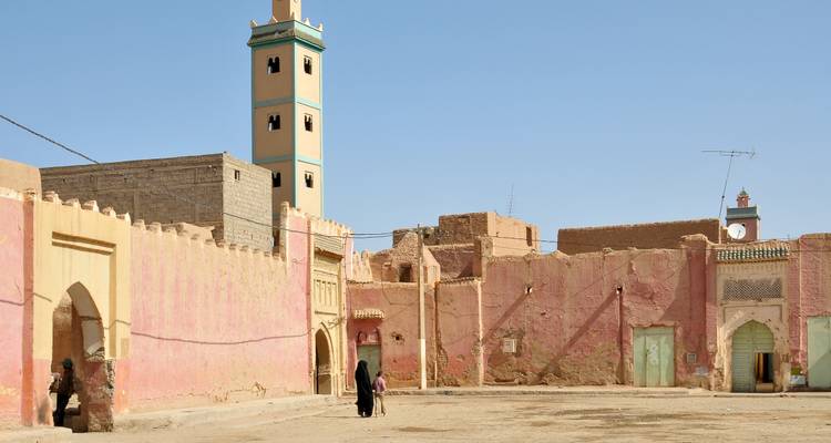 Sun-bleached pink walls and tall minaret tower framing an open dusty square with a few locals walking.