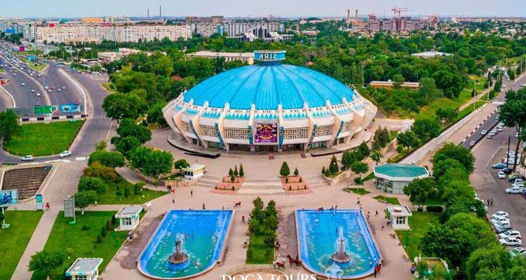 Vue aérienne de jour du Cirque de Tachkent avec dôme bleu, fontaines et avenues environnantes, filigrane présent.