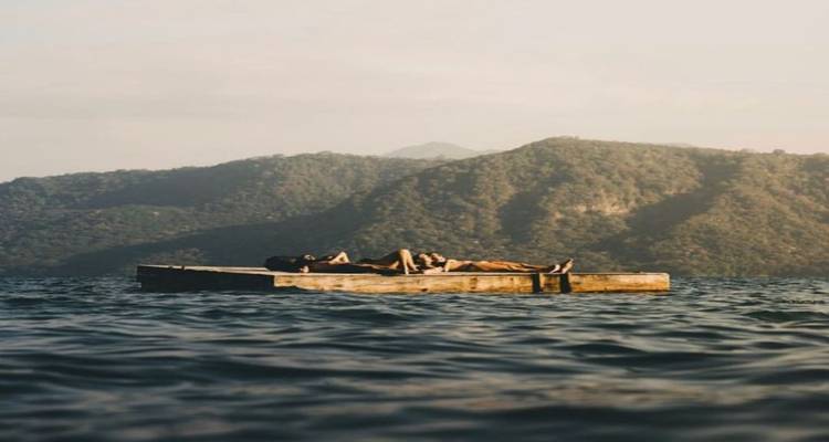 Deux voyageurs se détendent sur un petit radeau en bois flottant sur un lac tranquille entouré de collines boisées.