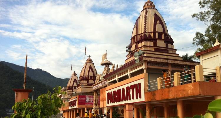 Flussufer Parmarth Niketan Ashram-Komplex mit cremefarbenen und kastanienbraunen Schreinen unter teilweise bewölktem Himmel