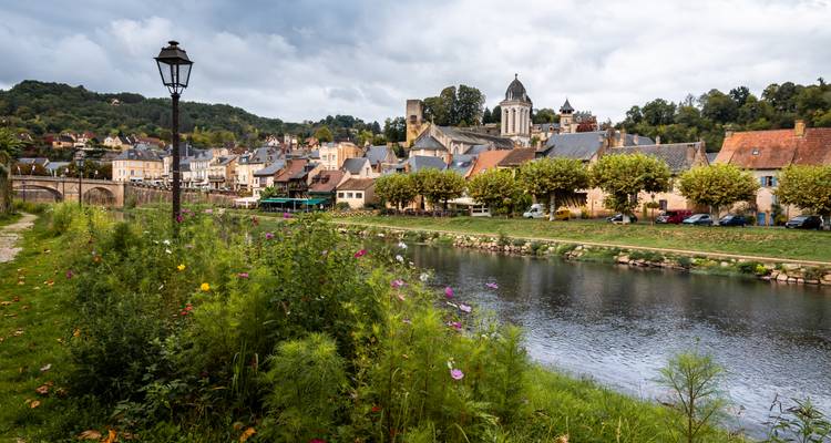 Ein charmantes französisches Dorf mit Steinhäusern und einem Flussuferweg, gesäumt von Wildblumen unter einem stimmungsvollen Himmel.